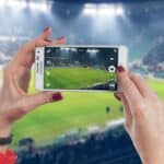Czerwony sweter i czarny zegarek na ręku, osoba robi zdjęcie meczu piłkarskiego na stadionie.